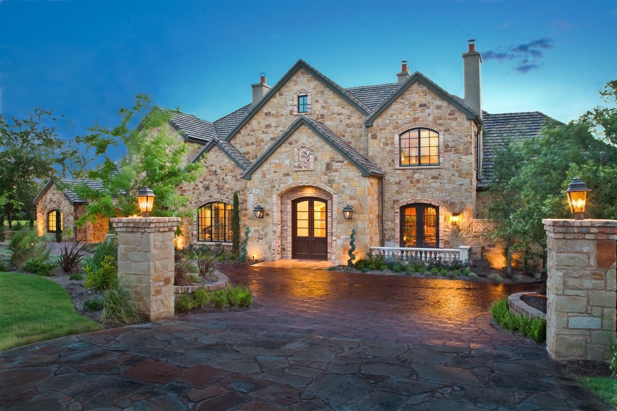 Stone house with arched front door, two chimneys, and a brick driveway lit by lamps.
