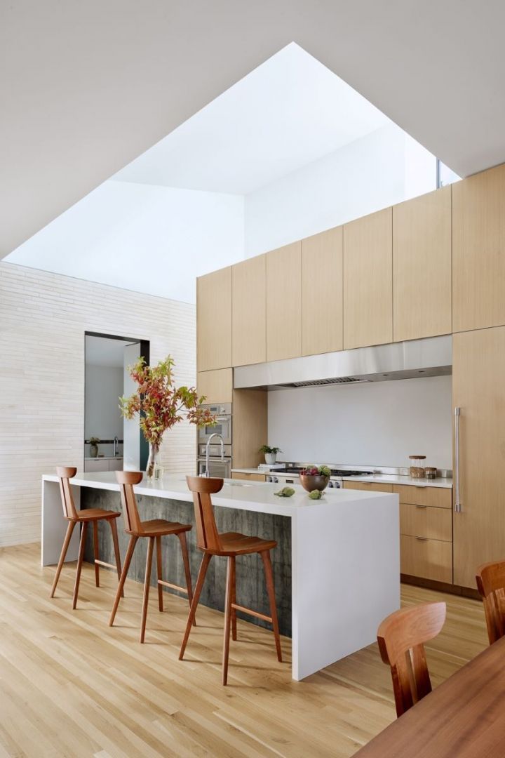 Modern kitchen with light wood cabinetry, white countertops, and wood stools. Natural light floods the space.