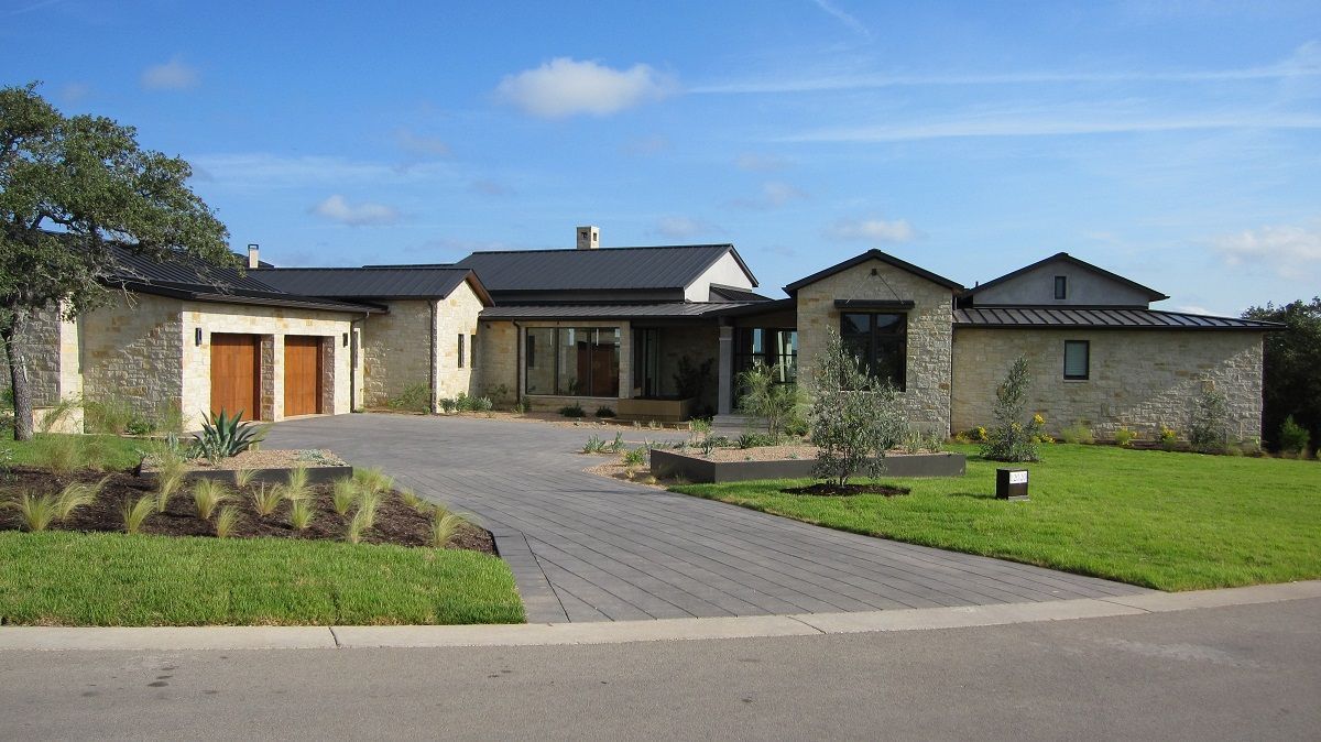 Stone house with dark roof, light brown garage doors, and a gray brick driveway on a sunny day.