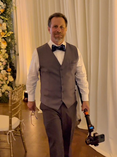A person in a gray vest and bowtie, holding a camera stabilizer, stands in a room with white drapes and floral decor.