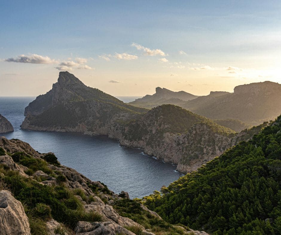 Paisaje costero de montañas con vistas a una bahía tranquila bajo un cielo parcialmente nublado al atardecer.