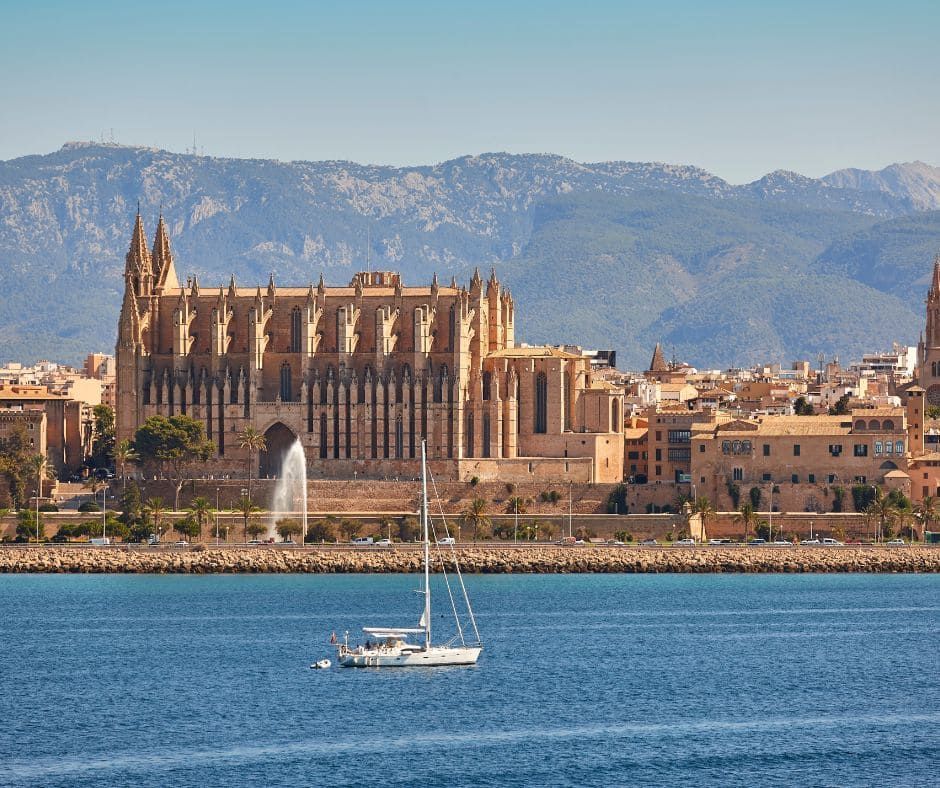 Catedral con vistas a un mar azul, con un velero, edificios y montañas al fondo en un día soleado.