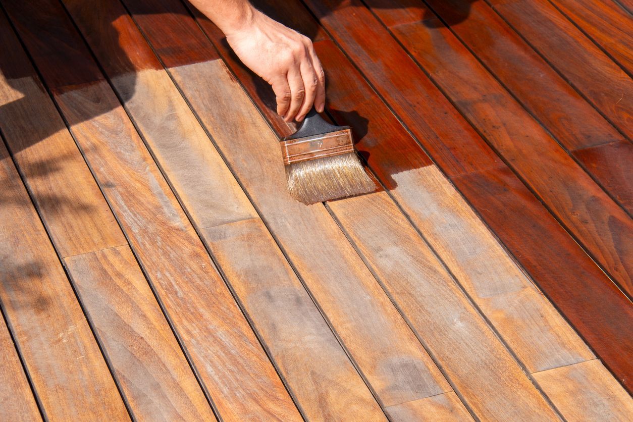 Person staining a wooden deck with a brush; section is newly stained dark brown.