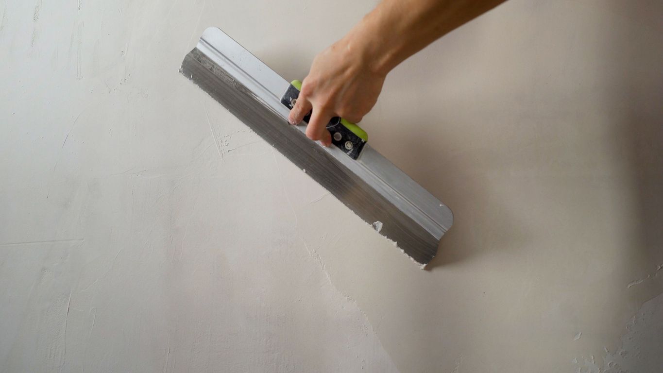 Hand using a trowel to smooth plaster on a white wall.