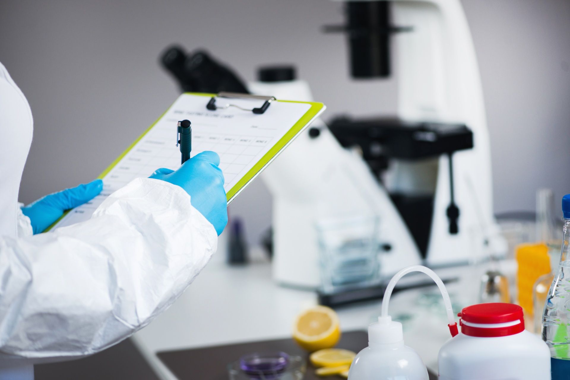 In un laboratorio luminoso, un tecnico di laboratorio con i guanti tiene in mano un blocco appunti accanto a un microscopio.