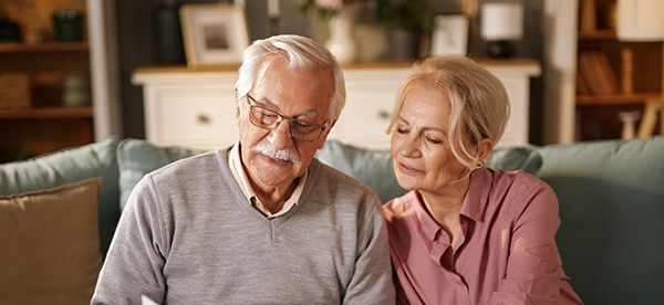 An elderly couple reads a document while seated on a teal couch indoors. Woman rests hand on man's shoulder.