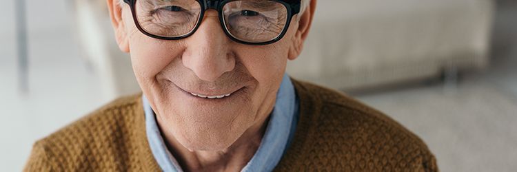 An elderly man wearing glasses and a brown sweater is smiling.