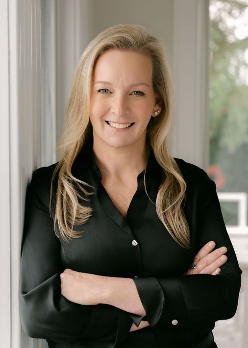 Woman in black shirt, arms crossed, smiling. Leans against a white wall near a window.