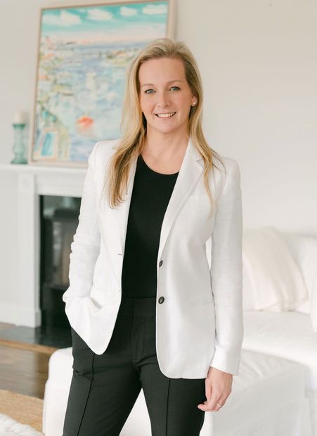 Woman in white blazer, black top and pants, smiling. Standing in living room with art.
