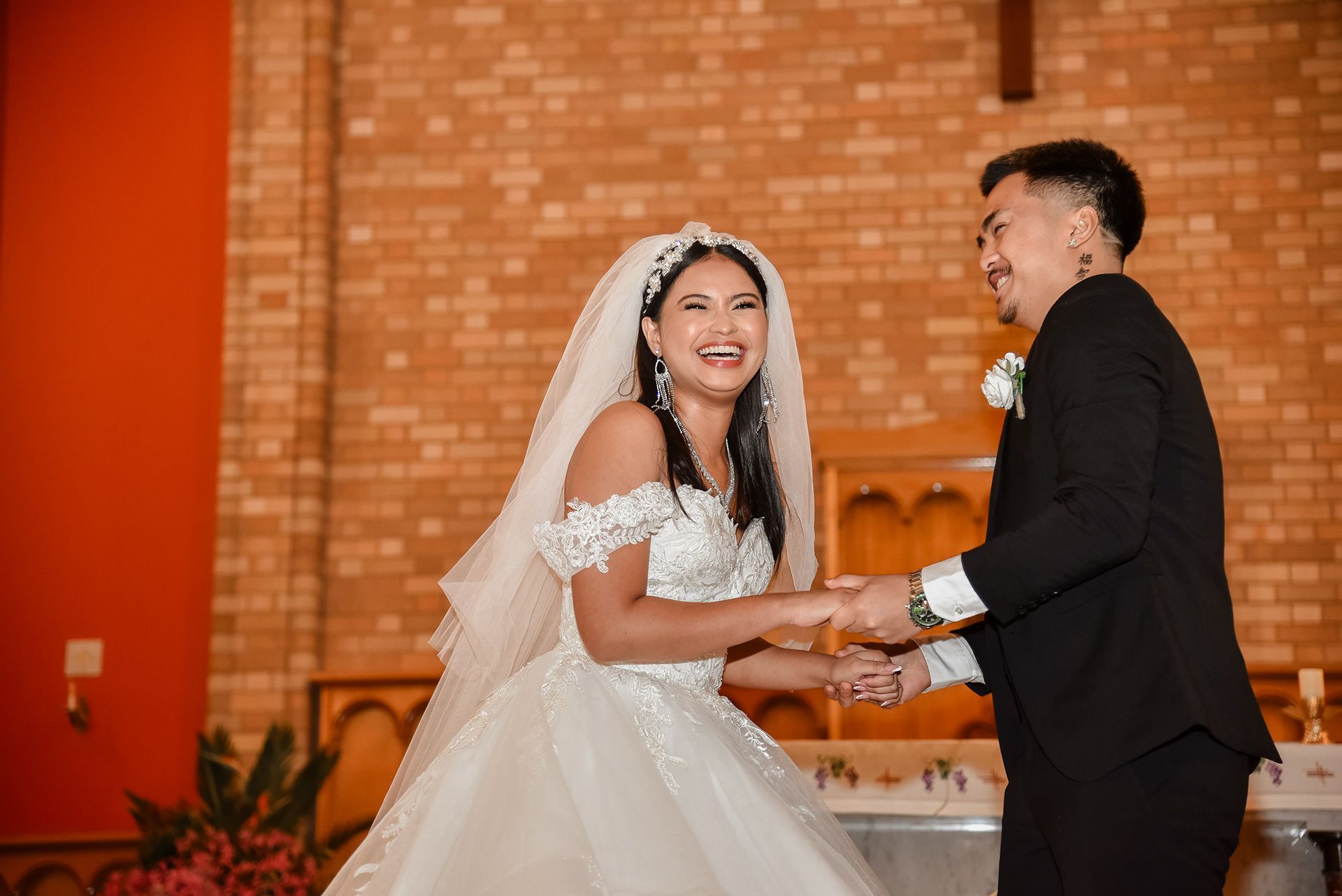 Bride and groom laughing, holding hands, indoors, wedding attire, brick wall backdrop.
