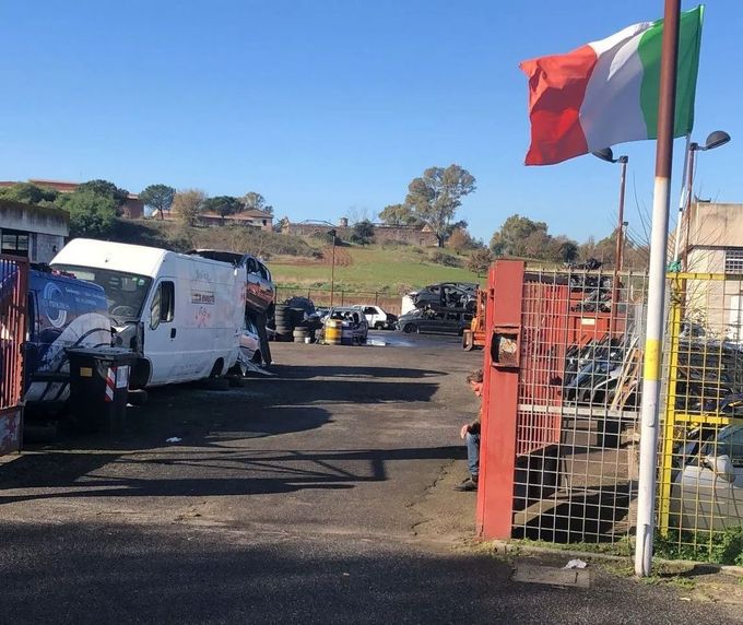 Parcheggio esterno con furgoni e automobili accanto a un cancello rosso e una bandiera in una giornata di sole.