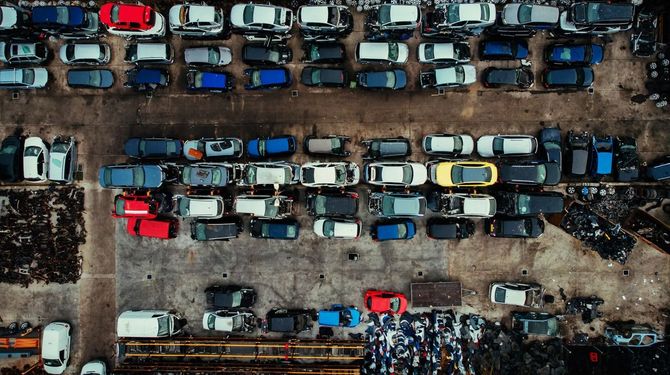 Veduta aerea di auto e furgoni parcheggiati uno accanto all'altro in un parcheggio fangoso, con file di veicoli blu, bianchi e rossi.