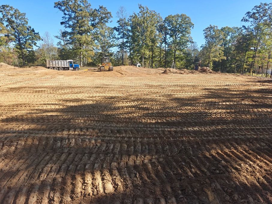 Cleared dirt field with tire tracks, trees along the edge, and a blue truck in the distance under a sunny sky