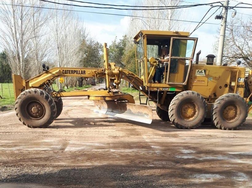 Una motoniveladora amarilla Caterpillar estacionada en un camino de tierra, con árboles y líneas eléctricas al fondo