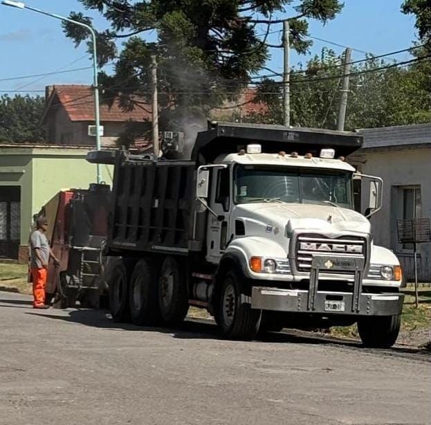 Tres obreros de la construcción con cascos inspeccionan una zona de obras viales, con un trípode de topografía y una excavadora al fondo.