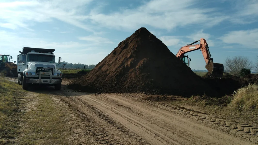 Camión volquete, excavadora y gran montón de tierra en una obra rural bajo un cielo despejado.