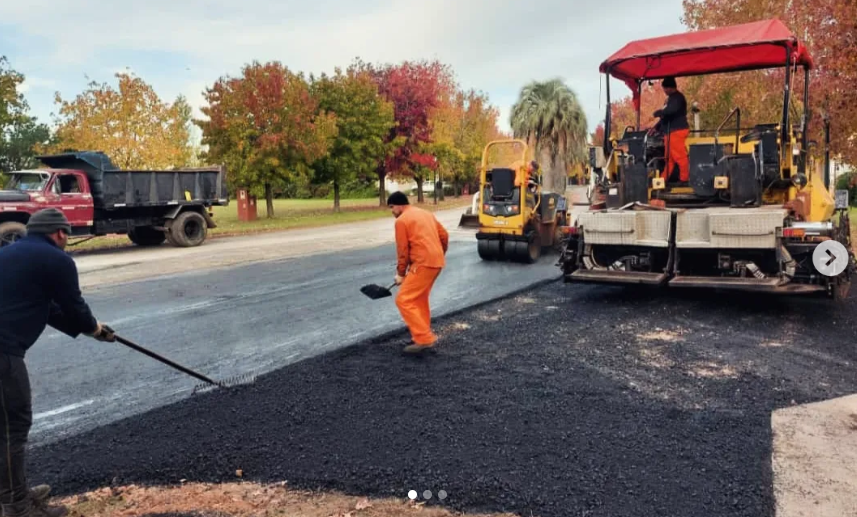 Equipo de construcción de carreteras pavimentando una calle con una extendedora de asfalto y trabajadores con equipo de seguridad naranja.