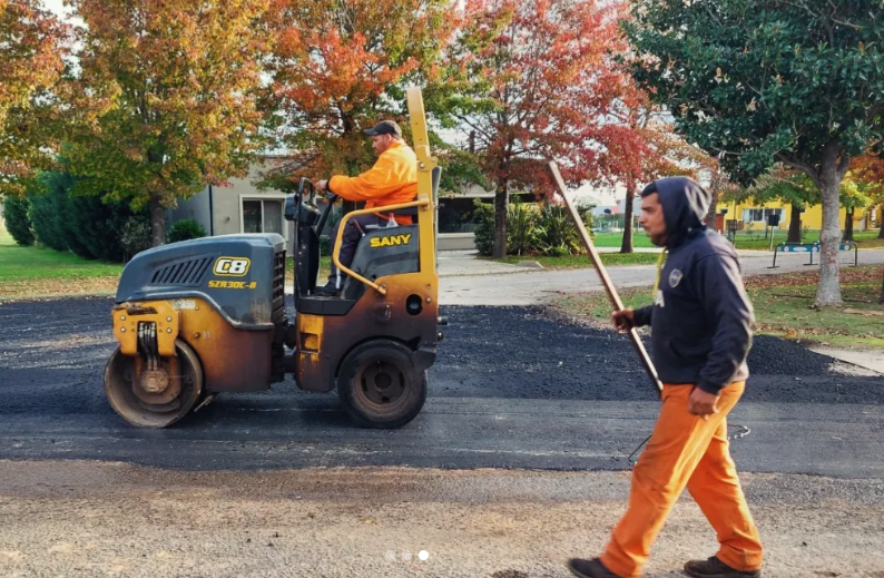 Dos trabajadores en una apisonadora compacta pavimentando un camino de entrada junto a árboles otoñales.