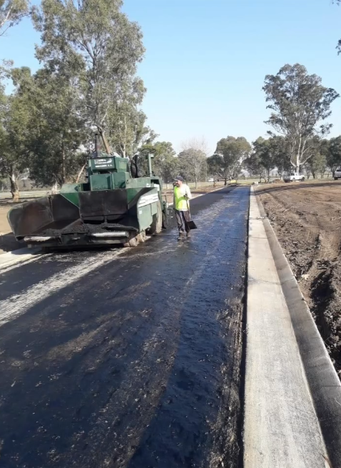 Un equipo de operarios viales está pavimentando un carril de asfalto negro junto a un bordillo de hormigón con una máquina pavimentadora verde.
