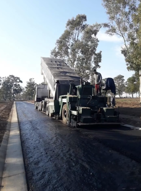 Máquina pavimentadora extendiendo asfalto nuevo en una carretera rural con trabajadores cerca.