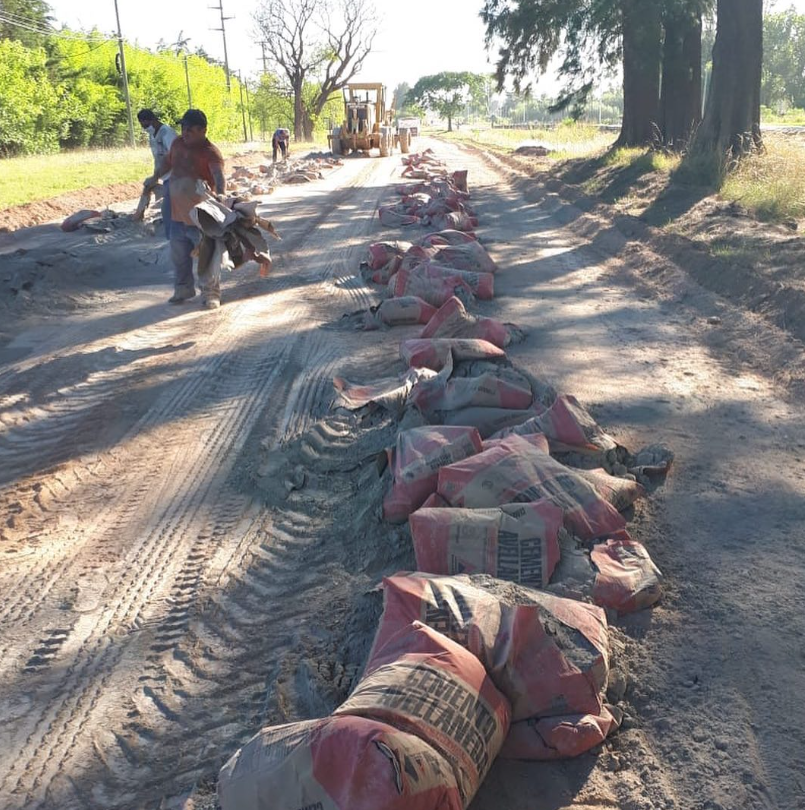 Sacos de tierra o grava apilados a lo largo de un camino polvoriento, con trabajadores y una cargadora al fondo.