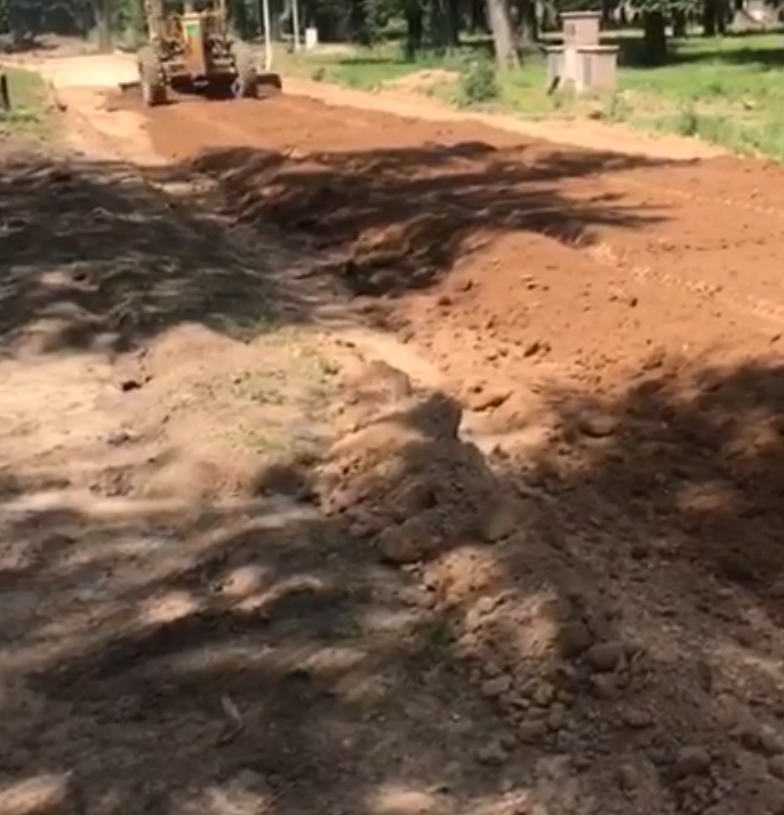Camino de tierra en construcción con una niveladora alisando la superficie en una zona boscosa soleada.