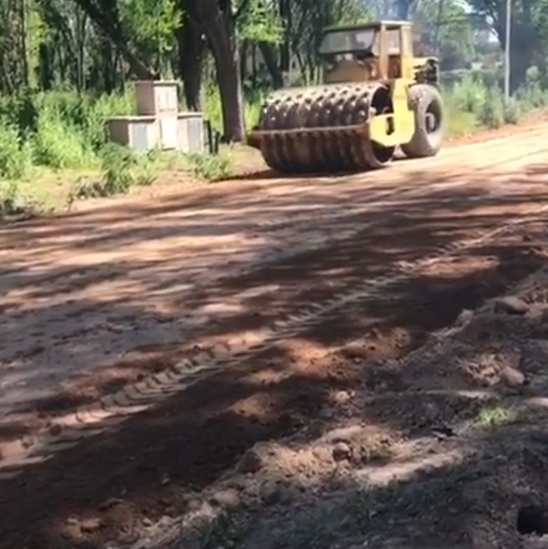Una apisonadora amarilla compacta un camino de tierra fangoso junto a árboles y un pequeño edificio.