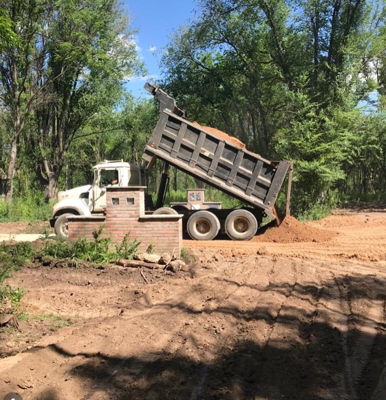 Un camión volquete blanco volcado en un terreno baldío con árboles al fondo.