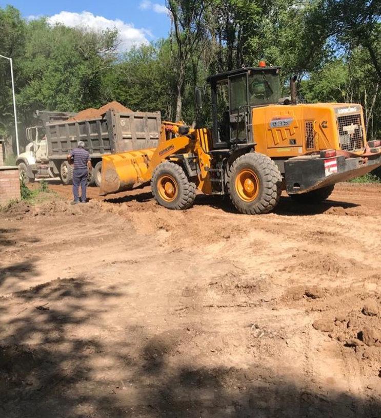 Una cargadora amarilla mueve tierra en una obra en construcción embarrada, con un trabajador cerca y árboles al fondo.