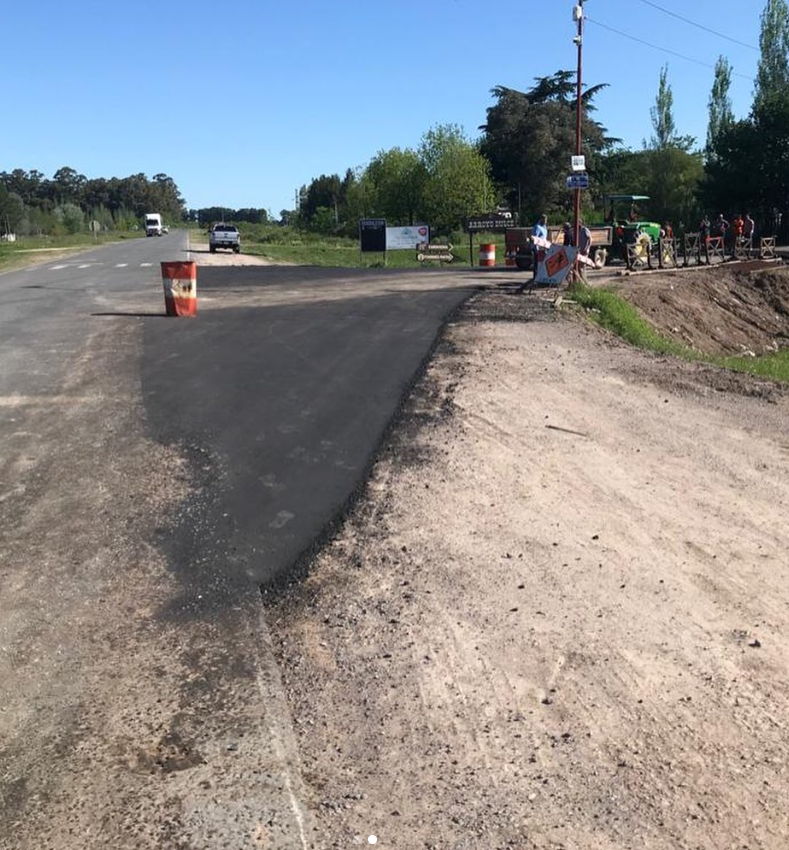 Carretera recién pavimentada junto a un arcén de tierra, con trabajadores y maquinaria cerca de la calzada bajo un cielo despejado.