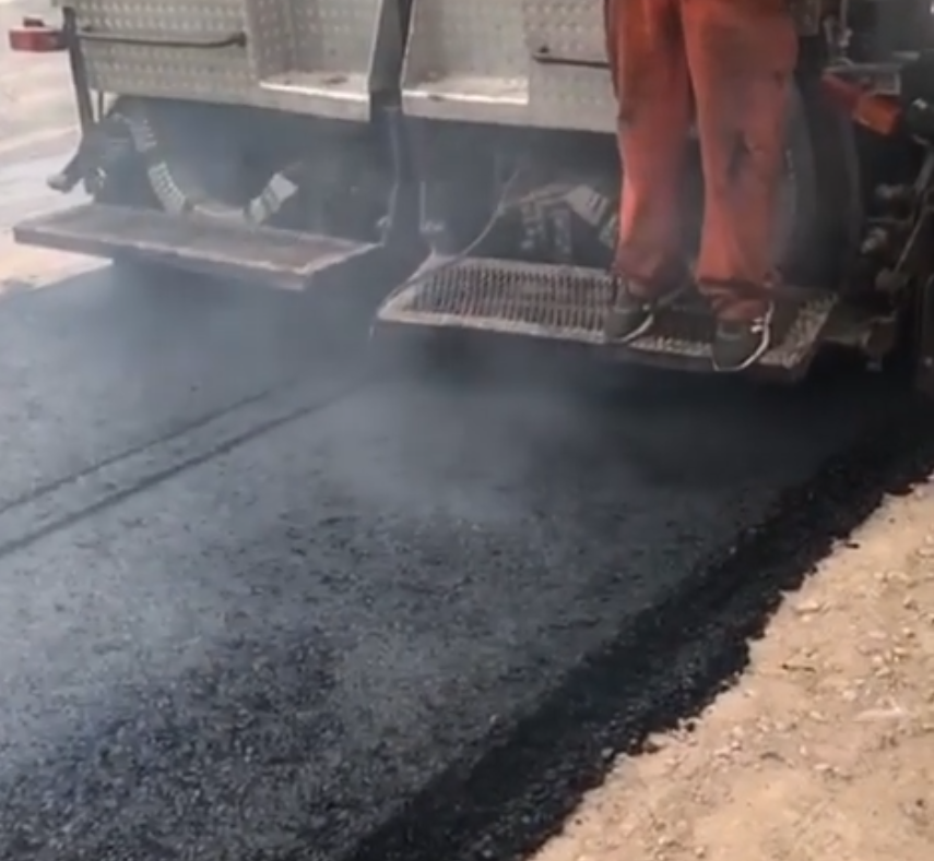Máquina pavimentadora extendiendo asfalto negro fresco junto a trabajadores con pantalones de seguridad naranjas.