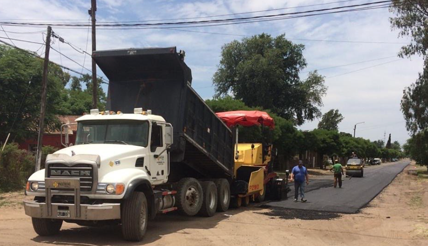 Camión volquete blanco con plataforma negra elevada estacionado al costado de la carretera, con un camión amarillo y trabajadores cerca bajo un cielo nublado.