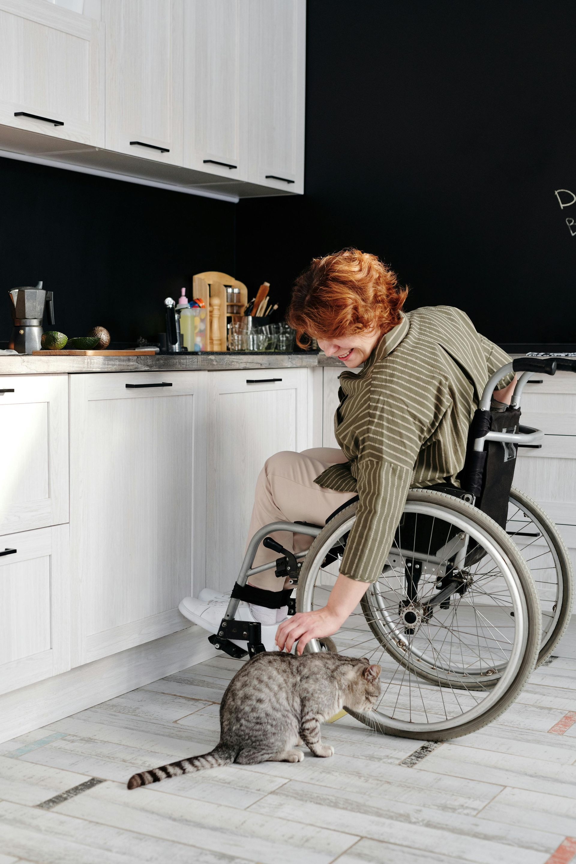 Woman in a wheelchair petting a gray tabby cat in a kitchen with white cabinets.