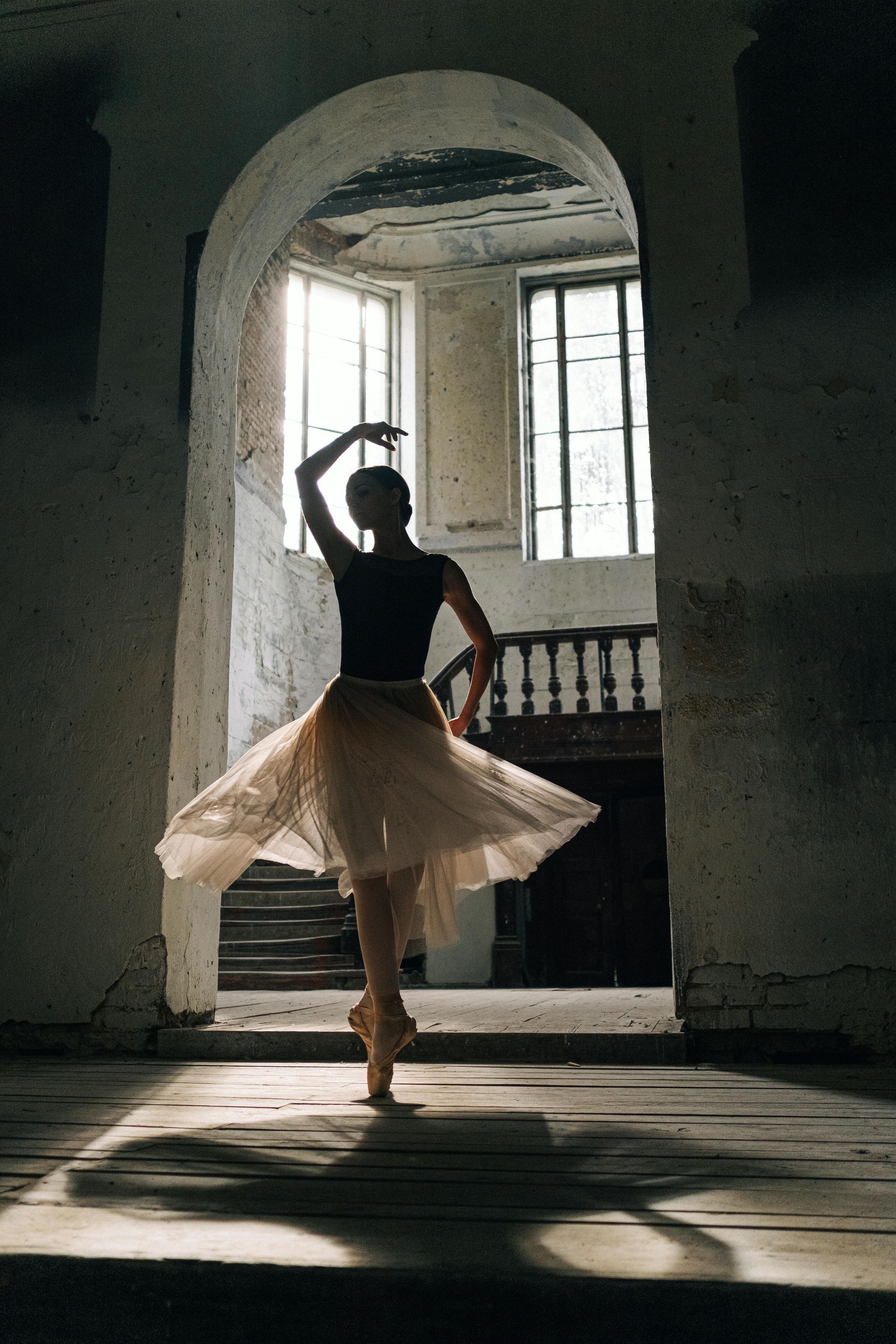 Ballerina in a tutu, silhouetted in an archway, on pointe. Light streams in from windows in an aged building.