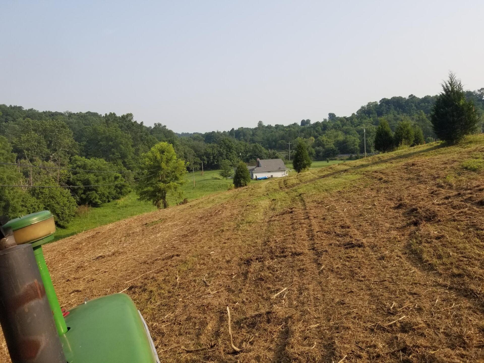 View from a tractor cab, looking down a freshly plowed field towards a house and tree-lined hills.