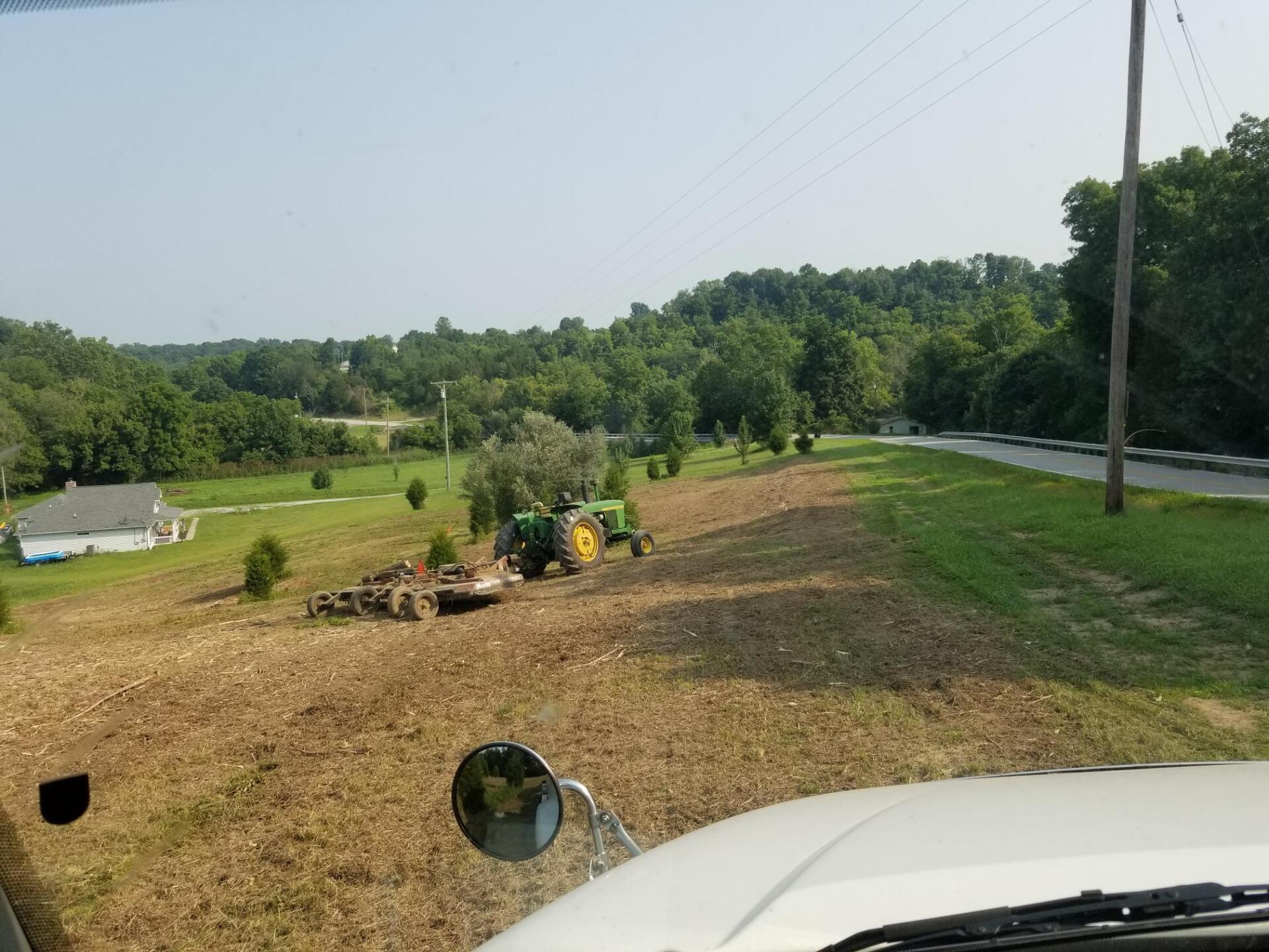 Tractor mowing a field next to a road, house and trees on a sunny day.