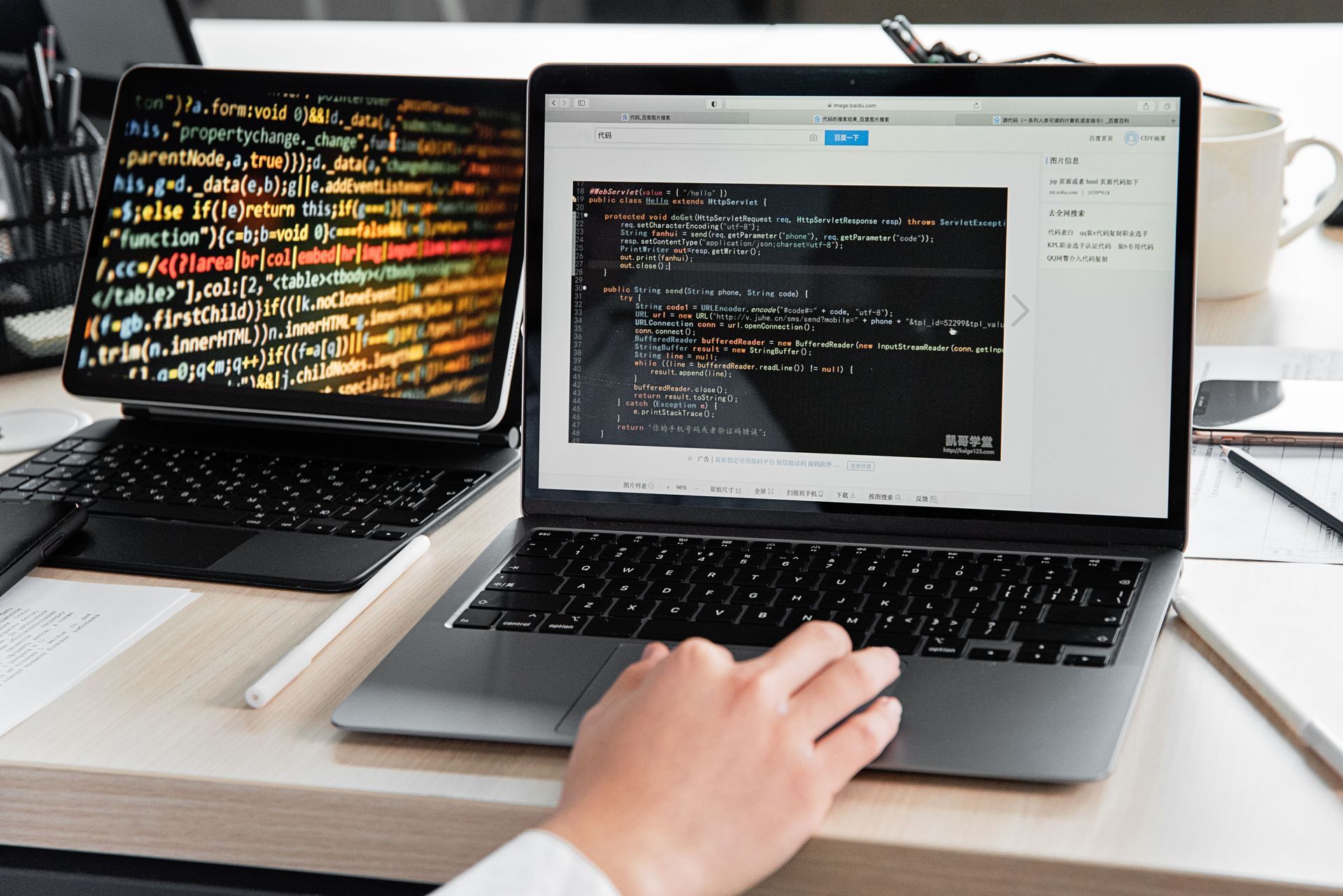 Two laptops on a desk displaying code, with a hand using the front laptop and a coffee mug nearby