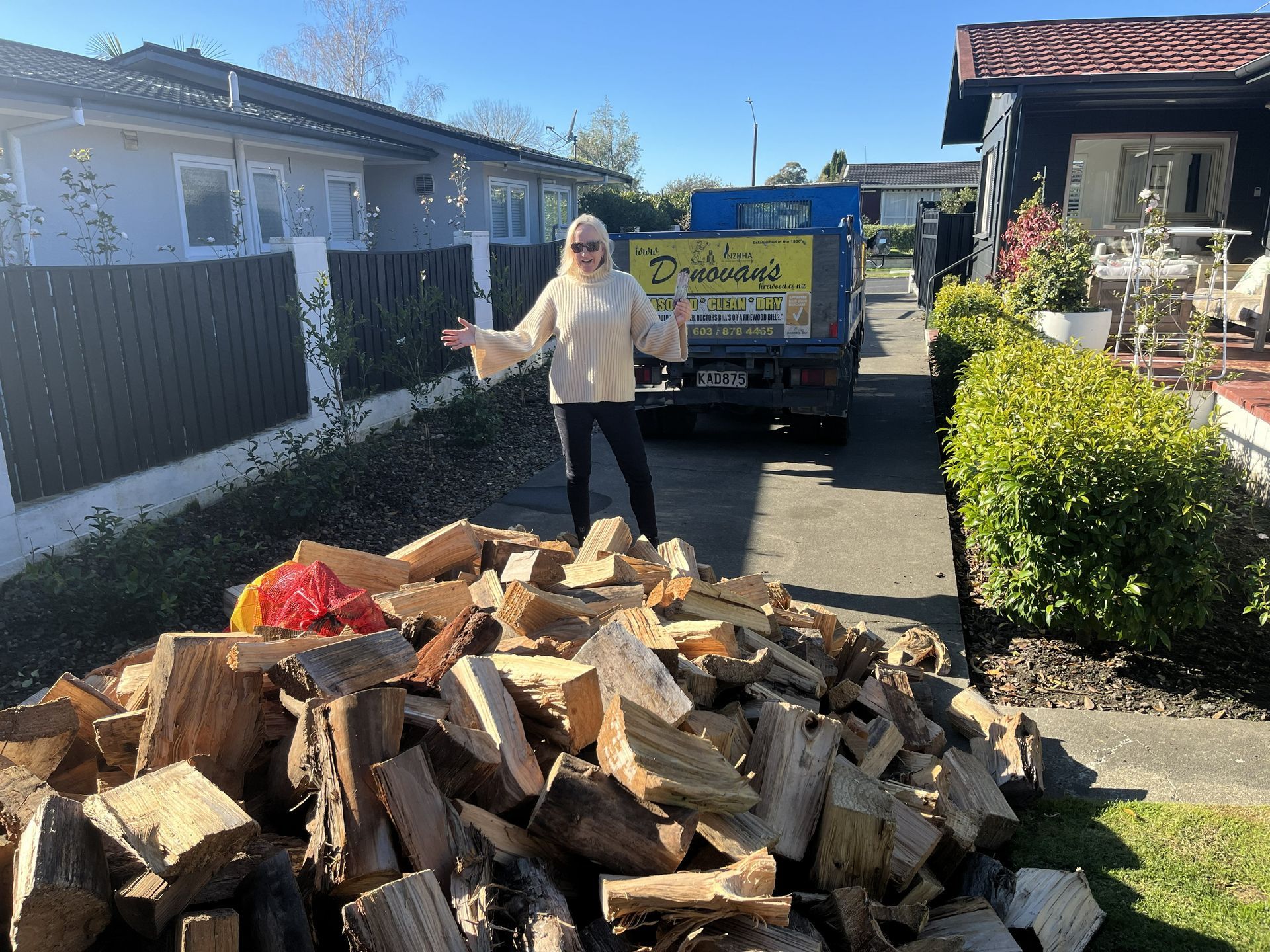 Person standing beside a large pile of cut firewood in a driveway, with a dump truck behind.