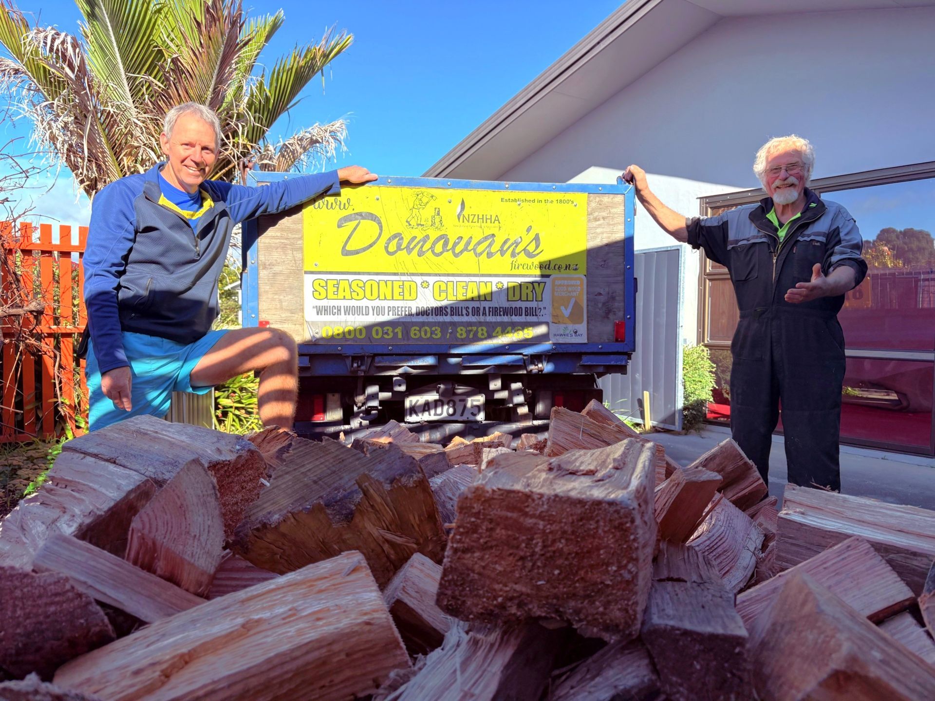Two men stand beside a truckload of firewood, one points at the pile. Sign reads 