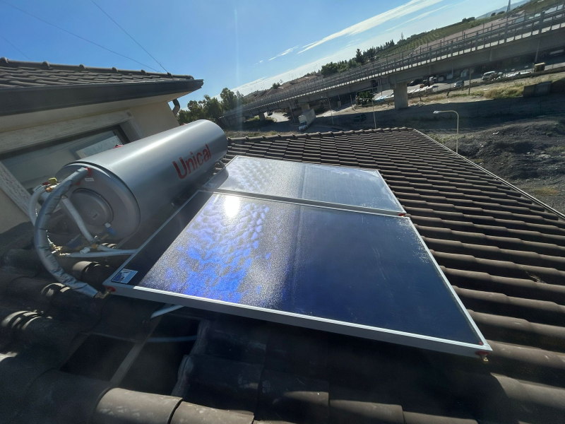 Scaldabagno solare su un tetto di tegole con serbatoio in acciaio inossidabile e pannelli solari blu; sullo sfondo un cavalcavia autostradale.