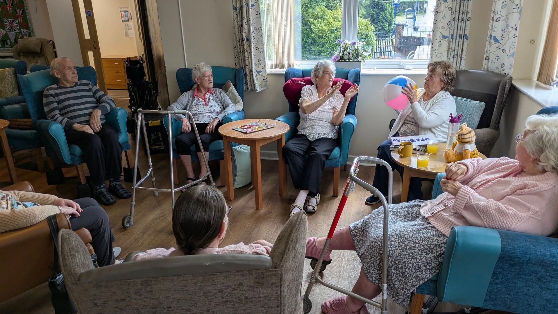 Residents sit in a cozy lounge, chatting in armchairs around a small table near a bright window.