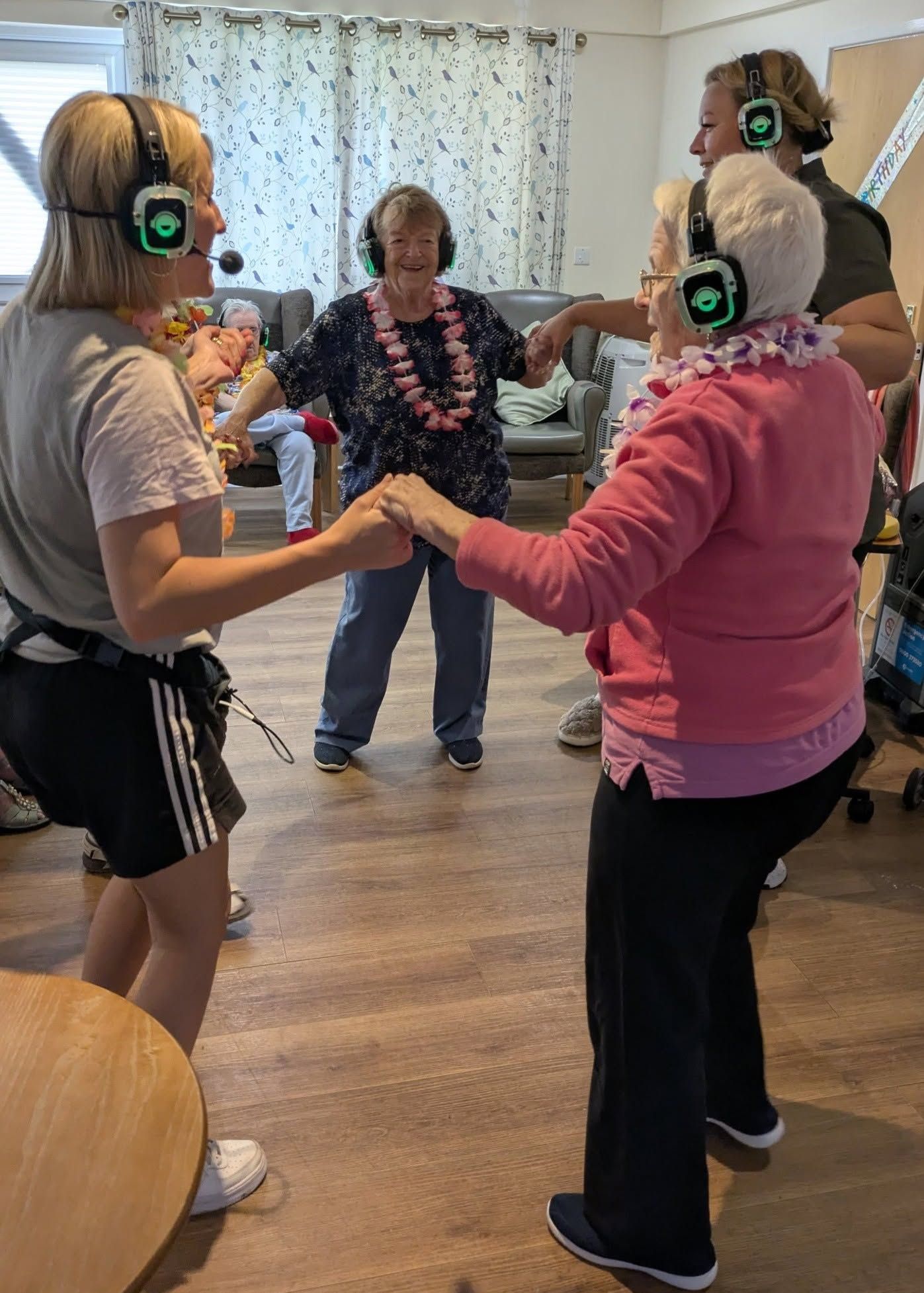 Three people wearing headphones stand in a circle holding hands while a woman leads them in a living room.