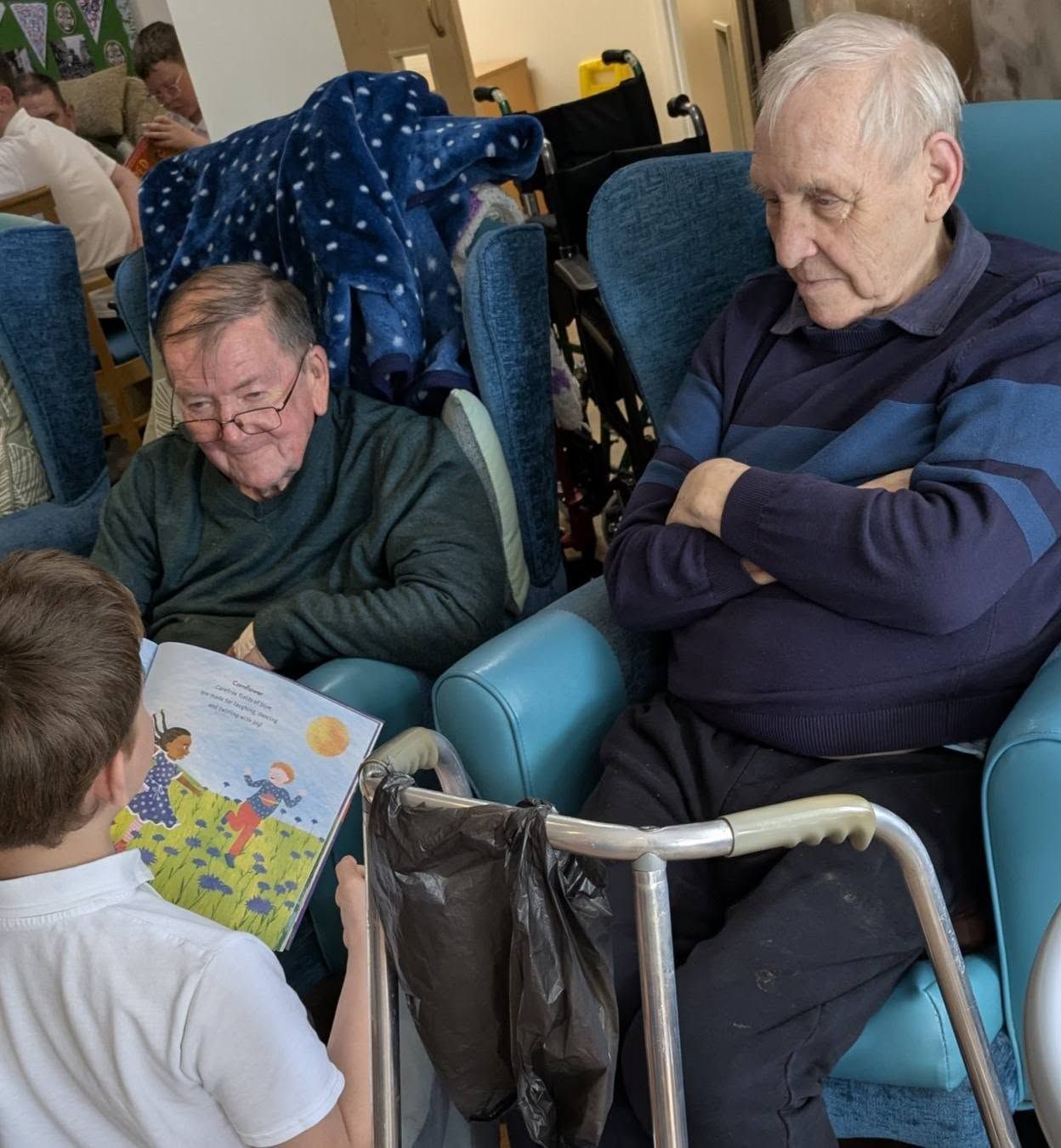 Two elderly men sit in armchairs while a child shows them a colorful book in a cozy room.