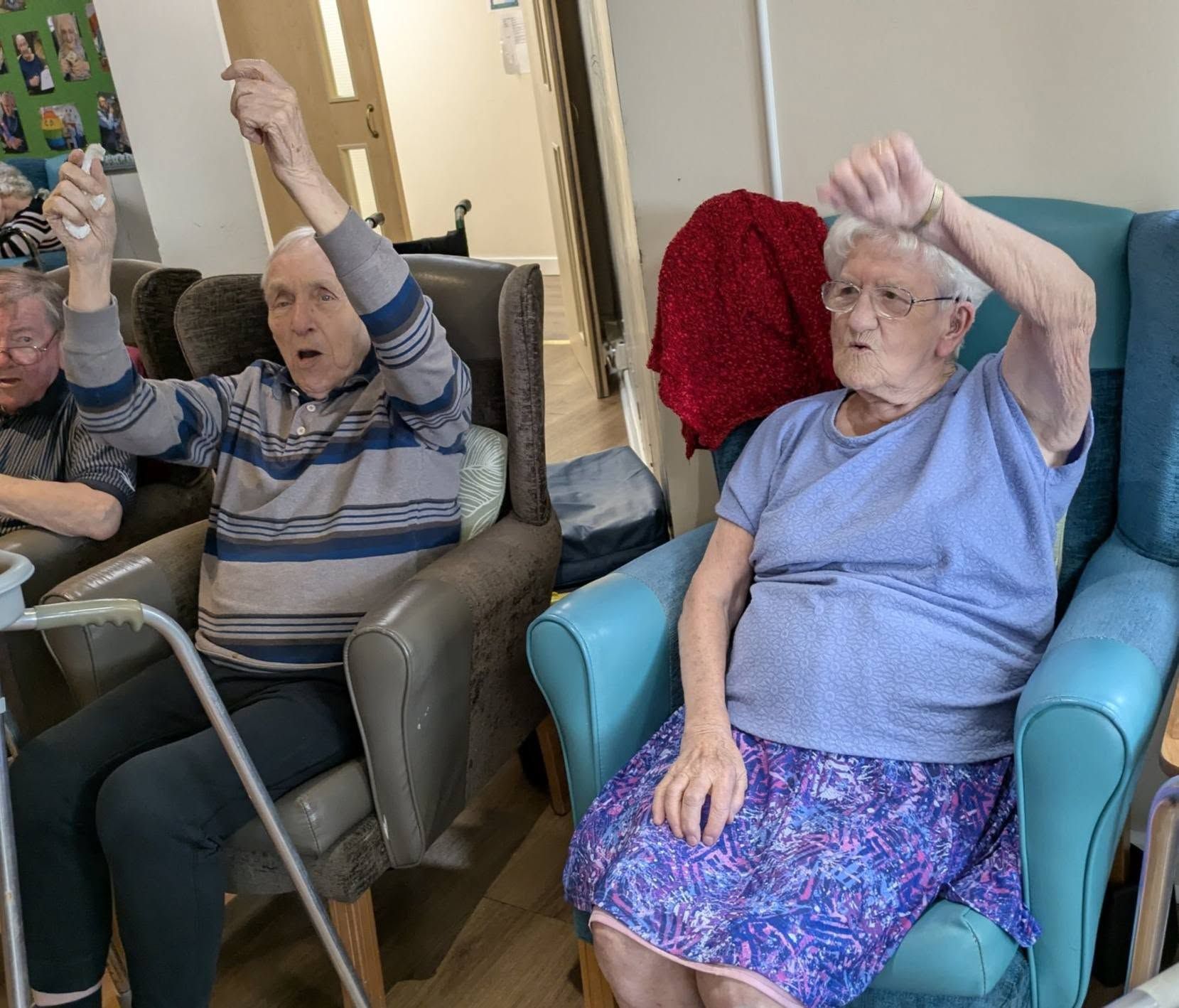 Two elderly people in armchairs raising their arms during a seated exercise session