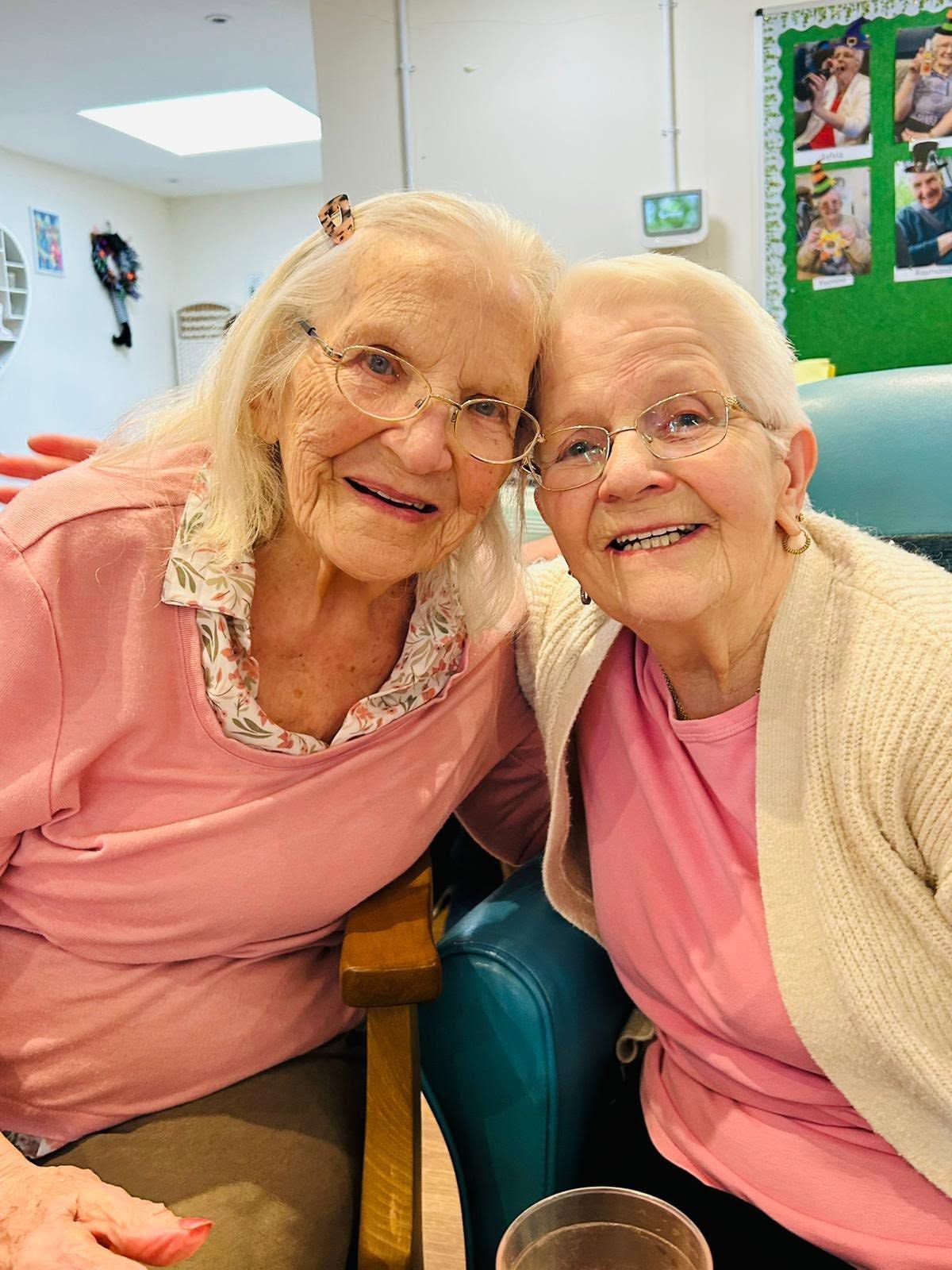 Two smiling elderly women sitting close together in a bright room, wearing pink tops and glasses.