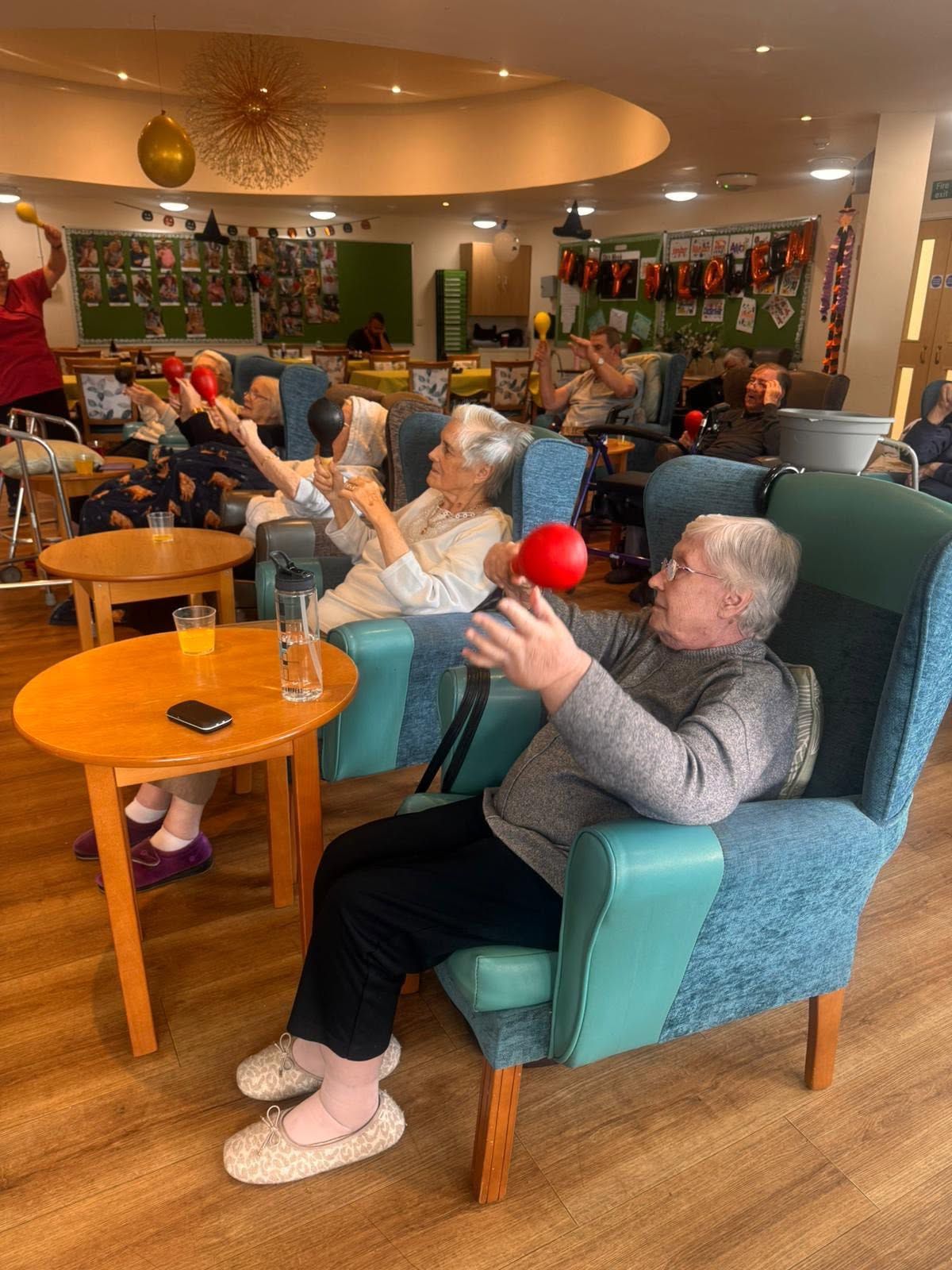 Residents doing seated exercise with red balls in a cozy communal lounge.