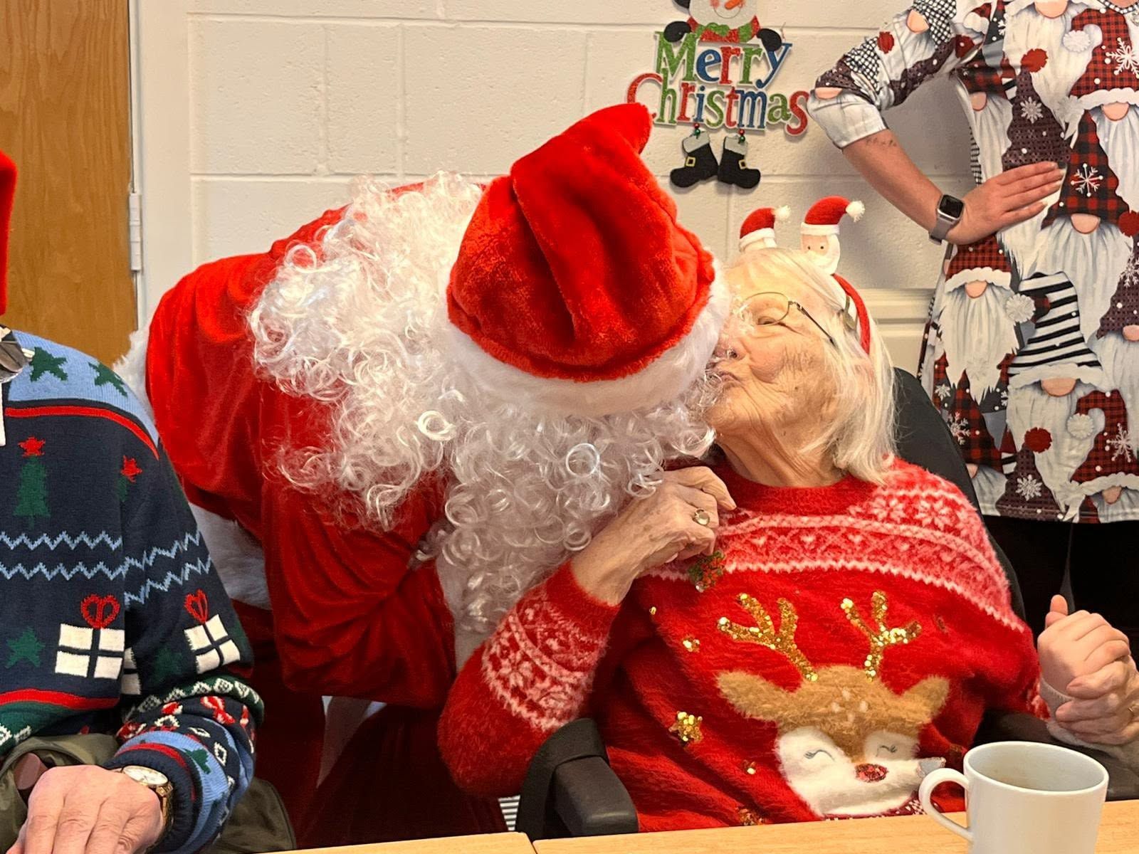 Santa leaning in to kiss a woman in a red Christmas sweater at a festive holiday gathering