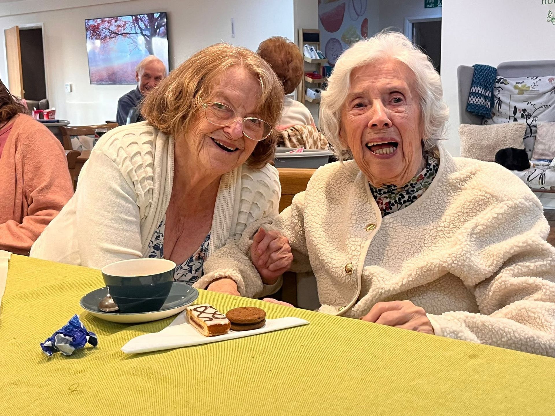 Two smiling women seated at a table with cups and pastries in a bright dining room