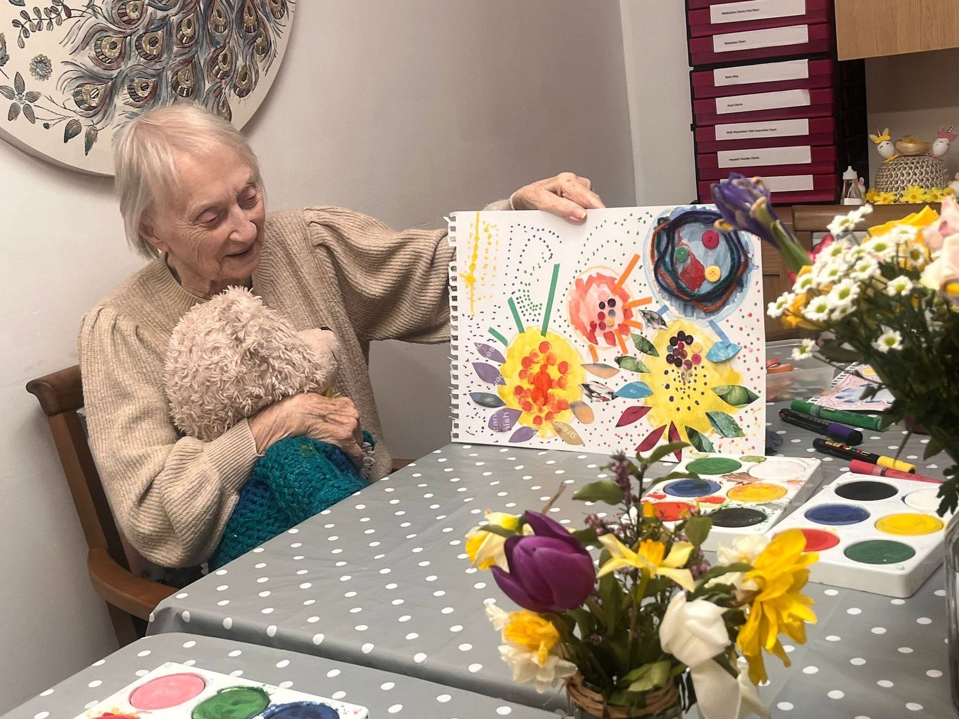Woman seated beside a child holding a colorful painting at a craft table with flowers and art supplies