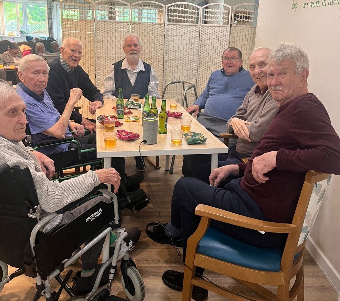 Group of older adults sharing a meal around a table in a bright dining room, one in a wheelchair